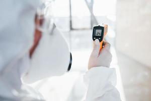 vrouw dokter wetenschapper in laboratorium jas, defensief eyewear en masker staand binnenshuis met geïnfrateerd thermometer foto