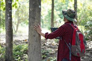 Aziatisch Mens botanicus draagt hoed , rood plaid overhemd en rugzak, houdt slim tablet naar controleren en enquête bomen in Woud. concept, gebruik makend van slim apparaat technologie naar beheren milieu en Onderzoek bomen. foto