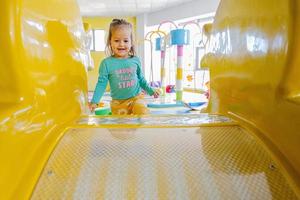 gelukkig meisje spelen Bij binnen- Speel centrum speelplaats. glijden in geel schuiven. foto