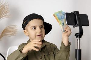 schattig, schattig, weinig jongen blogger opname levensstijl blog, Holding geld en pratend naar camera van smartphone Aan statief. jong influencer filmen vlog voor zijn kanaal. kind maakt video. foto