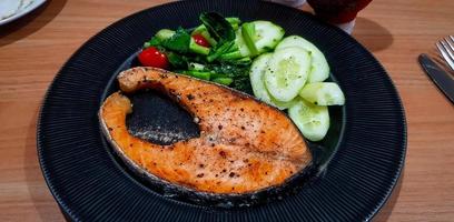 gegrild Zalm steak met plakjes komkommer, tomaat en vers groente Aan zwart schotel of bord Aan houten tafel voor lunch tijd. gezond voedsel en leven stijl aan het eten. vis met topping zwart peper en zout foto