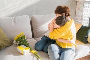 moeder knuffelen haar zoon zittend Aan de bank. geschenk en bloemen in de buurt. verjaardag, moeder dag of vrouwen dag concept foto