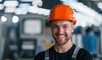 glimlachen en gelukkig medewerker. portret van industrieel arbeider binnenshuis in fabriek. jong technicus met oranje moeilijk hoed foto