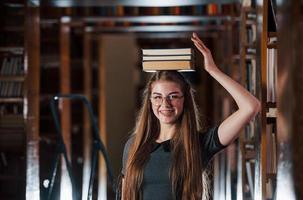 vrolijk meisje hebben pret terwijl zet boeken Aan haar hoofd. vrouw leerling is in bibliotheek. opvatting van onderwijs foto