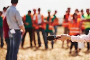 vrouw hand- houdt microfoon. groep van mensen in geel uniform staat buitenshuis Aan de veld- foto