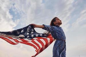 patriottisch vrouw kind met Amerikaans vlag in handen. tegen bewolkt lucht foto