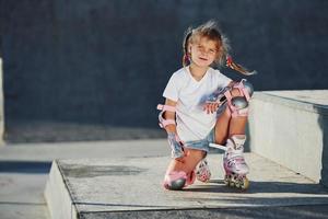 schattig weinig meisje met rol skates buitenshuis zit Aan de oprit voor extreem sport- foto