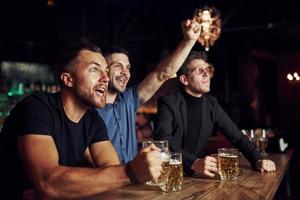 vieren zege. drie sport- fans in een bar aan het kijken voetbal. met bier in handen foto