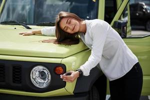 zij is in liefde met haar nieuw auto. sleutels in hand. jong vrouw in wit officieel kleren staat in voorkant van groen auto- buitenshuis foto