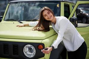 zij is in liefde met haar nieuw auto. sleutels in hand. jong vrouw in wit officieel kleren staat in voorkant van groen auto- buitenshuis foto