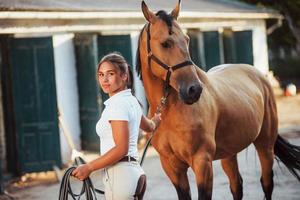 krijgen klaar voor de rijden. amazone in wit uniform met haar paard Bij boerderij. klaar voor de rijden foto