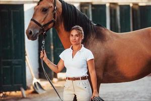 krijgen klaar voor de rijden. amazone in wit uniform met haar paard Bij boerderij. klaar voor de rijden foto