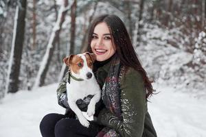 vrolijk meisje knuffels haar huisdier. glimlachen brunette hebben pret terwijl wandelen met haar hond in de winter park foto