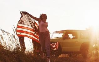 meisje loopt naar voren. vrienden hebben mooi hoor weekend buitenshuis in de buurt van hen groen auto met Verenigde Staten van Amerika vlag foto