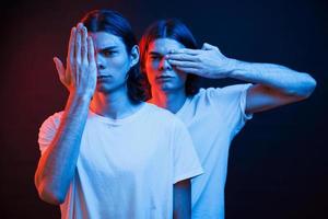 aan het bedekken ogen met handen. portret van tweeling broers. studio schot in donker studio met neon licht foto