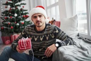 echt Look. foto van Mens in de kerstman hoed en vakantie kleren houdt nieuw jaar geschenk. Kerstmis boom Aan achtergrond