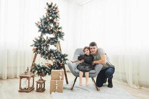 samen in de kamer. Kerstmis en vakantie conceptie. schattig weinig meisje met haar vader is zit Aan de stoel in de buurt ladder versierd met sterren en geschenk dozen Aan de verdieping foto