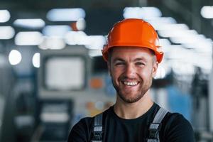 glimlachen en gelukkig medewerker. portret van industrieel arbeider binnenshuis in fabriek. jong technicus met oranje moeilijk hoed foto
