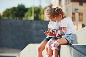 gebruik makend van smartphone. Aan de oprit voor extreem sport. twee weinig meisjes met rol skates buitenshuis hebben pret foto