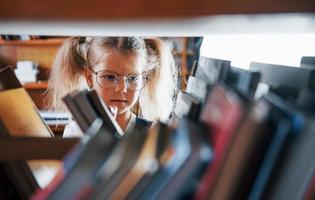 weinig meisje in bril zoeken voor een boek in de bibliotheek. opvatting van onderwijs foto