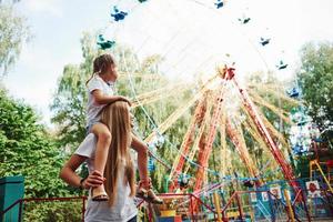 dochter zit Aan de schouders. vrolijk weinig meisje haar moeder hebben een mooi zo tijd in de park samen in de buurt attracties foto