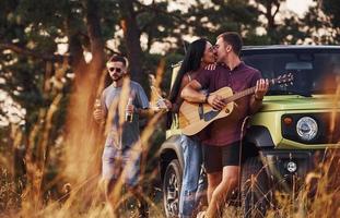 spelen Aan akoestisch gitaar en zingen. groep van vrolijk vrienden hebben mooi hoor weekend Bij zonnig dag in de buurt van hen groen auto buitenshuis foto