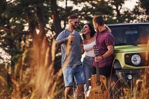 Mens houdt gitaar. groep van vrolijk vrienden hebben mooi hoor weekend Bij zonnig dag in de buurt van hen groen auto buitenshuis foto