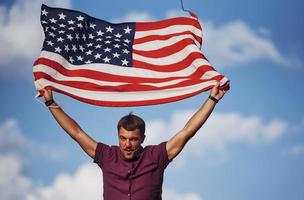 patriottisch gelukkig Mens golvend Amerikaans vlag tegen bewolkt blauw lucht foto