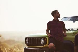 portret van Mens dat leunend Aan groen auto Bij mooi zonnig dag buitenshuis in de veld- foto