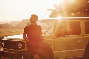 portret van Mens dat leunend Aan groen auto Bij mooi zonnig dag buitenshuis in de veld- foto