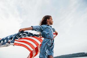 patriottisch vrouw kind met Amerikaans vlag in handen. tegen bewolkt lucht foto