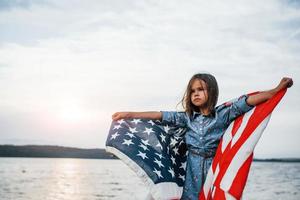patriottisch vrouw kind met Amerikaans vlag in handen. tegen bewolkt lucht foto