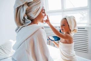 gebruik makend van room naar Doorzichtig huid. jong moeder met haar dochter hebben schoonheid dag binnenshuis in wit kamer foto