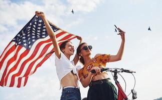 tegen bewolkt lucht. twee patriottisch vrolijk vrouw met fiets en Verenigde Staten van Amerika vlag in handen maakt selfie foto