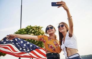 groen boom Bij achtergrond. twee patriottisch vrolijk Dames met Verenigde Staten van Amerika vlag in handen maken selfie buitenshuis in park foto