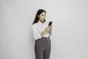 een portret van een gelukkig Aziatisch vrouw vervelend een wit overhemd en Holding haar telefoon, geïsoleerd door wit achtergrond foto