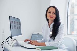 brunette vrouw dokter zit in modern kantoor door computer en looks Bij camera foto