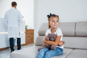 artsen achter. schattig weinig meisje met teddy beer in handen zit in aan het wachten kamer van ziekenhuis foto