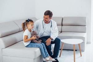 mannetje dokter in wit uniform zit in de kliniek met weinig meisje foto