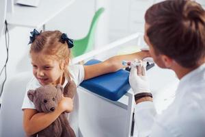 bloed bemonstering. weinig meisje met haar teddy beer is in de kliniek met dokter foto