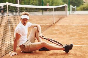 moe tennis speler in sportief kleren is Aan de rechtbank buitenshuis leunend Aan de netto foto