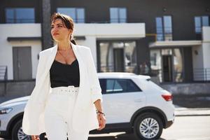 zelfverzekerd vrouw in wit formeel slijtage buiten in de buurt haar wit auto foto