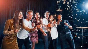 hebben pret met sterretjes. confetti is in de lucht. groep van vrolijk vrienden vieren nieuw jaar binnenshuis met drankjes in handen foto