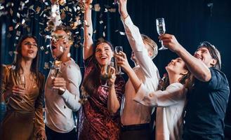 confetti is in de lucht. groep van vrolijk vrienden vieren nieuw jaar binnenshuis met drankjes in handen foto