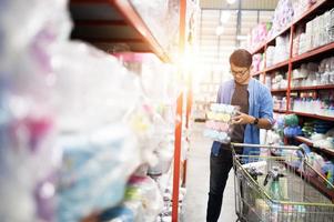 Aziatisch Mens winkelen. knap glimlachen jong Mens boodschappen doen voor huishouden items met supermarkt en boodschappen doen kar. foto