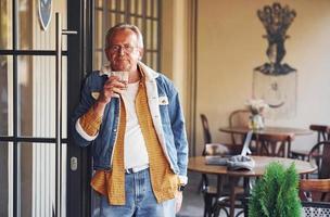 elegant senior in modieus kleren en in bril staat in de cafe met drinken in hand- foto