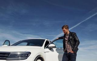 Mens in zwart leer jasje staat in de buurt zijn geparkeerd wit auto buitenshuis tegen blauw lucht foto