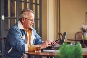 elegant senior in modieus kleren en in bril zit in de cafe en werken met laptop foto