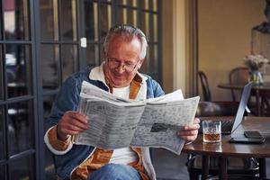 elegant senior in modieus kleren en in bril zit in de cafe en leest krant- foto
