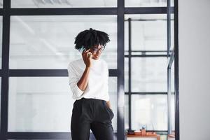 jong Afrikaanse Amerikaans vrouw in bril staat binnenshuis in de kantoor en hebben een telefoontje foto